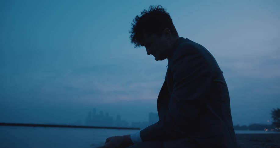 A distraught businessman sits on the beach sand at dusk