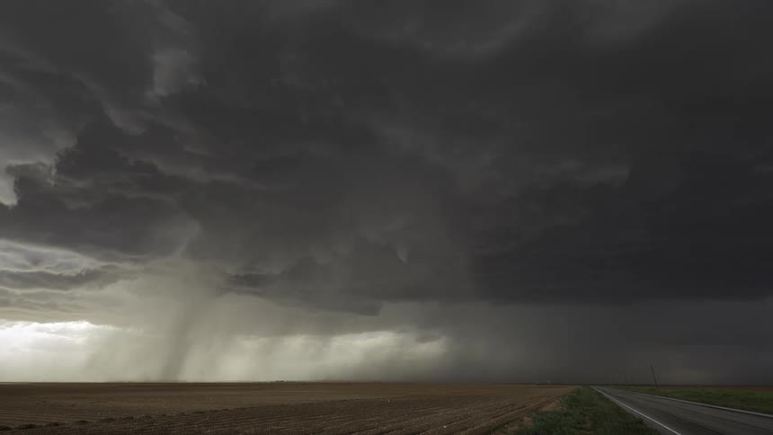 Rain falls over a field of flat farmland under swirling storm clouds as a truck drives by down the road
