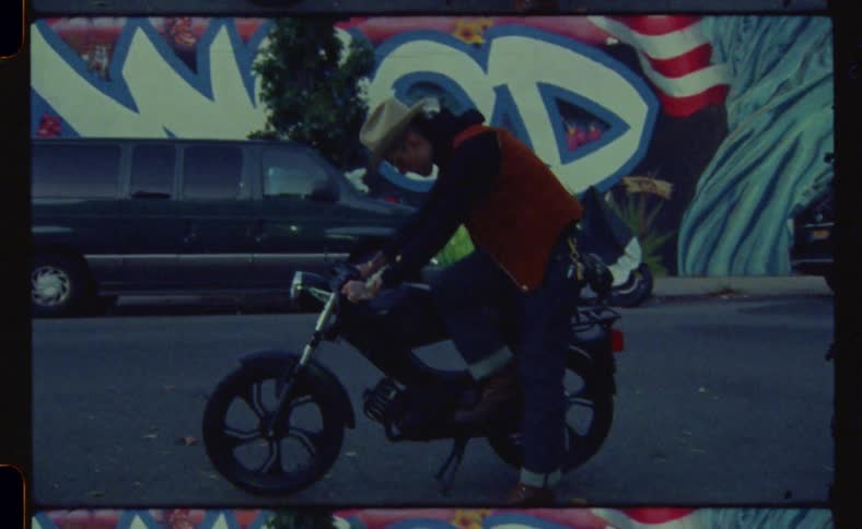 Man tries to kickstart a motorcycle in front of a wall covered in graffiti