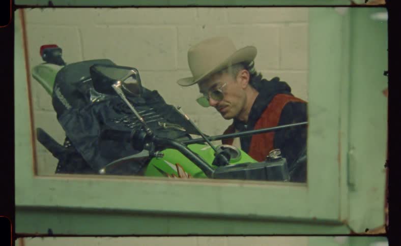 Man wearing sunglasses and a cowboy hat repairs a motorcycle on a small workshop