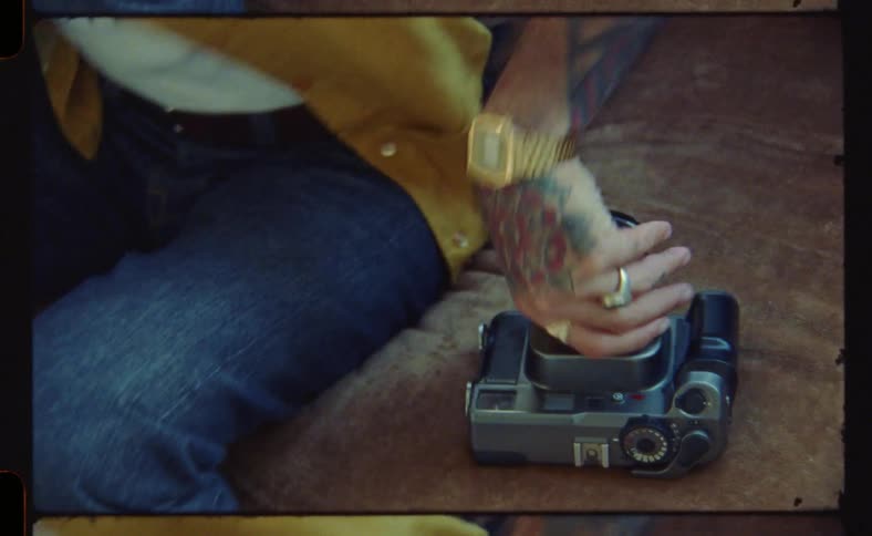 A man with tattooed arms and hands wearing jeans sits on a brown couch beside a camera grabs camera and stands up