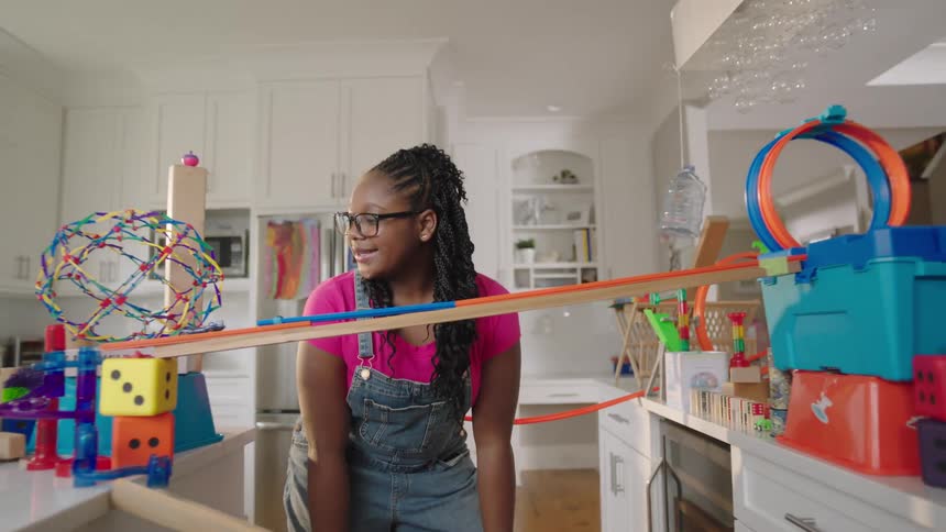 A young girl watching a toy car on a toy ramp in a kitchen during the day