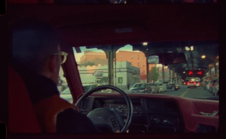 Man with glasses and red vest drives car down the street