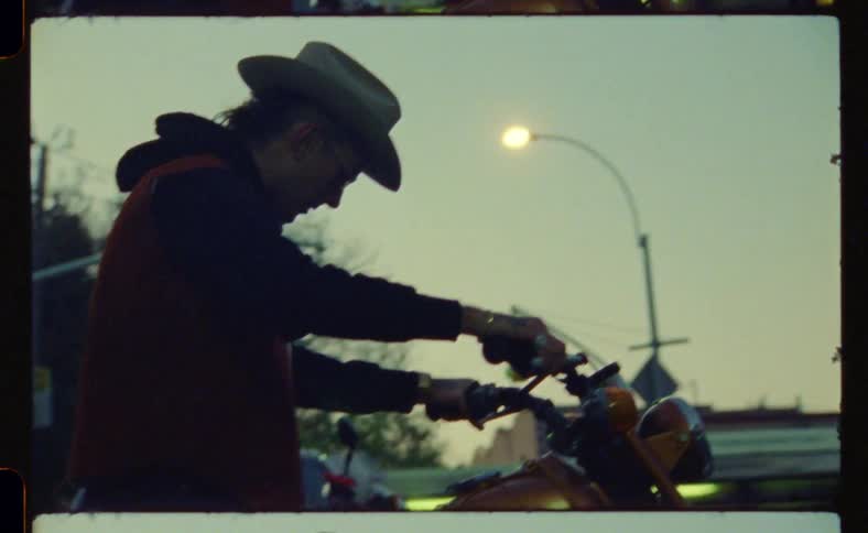 In the middle of the street a man wearing a hat and sunglasses struggles to start his bike a street light and a traffic sign stand in the background
