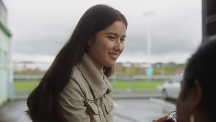 A woman sitting outdoors near a parking lot smiles while showing a friend her camera