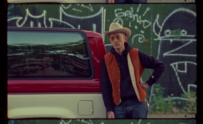A man wearing a cowboy hat is leaning against a car and looking around as if hes expecting someone