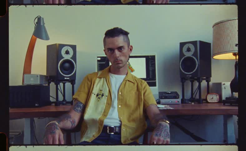 A man in a yellow shirt is sitting and looking forward and just behind him there is a computer with speakers