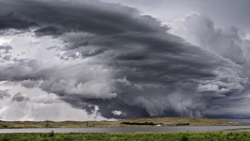 Time lapse of white and gray storm clouds rolling over low hills during the day with a small body of water