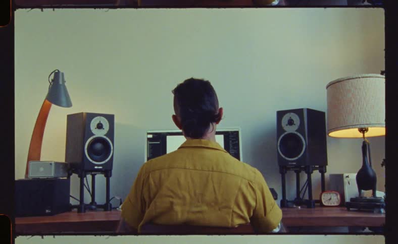 The backside of a man sitting at a desk in front of a computer speakers and lamp during the day