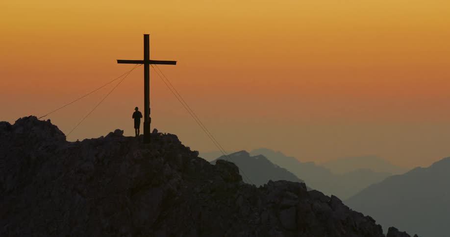 Person next to the cross on top of a mountain at the end of the day