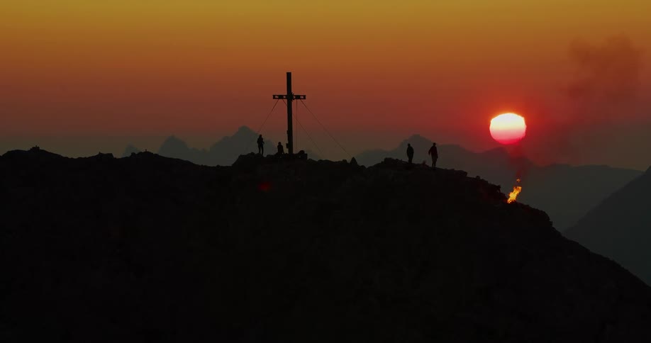 Silhouetted people on mountain topped with cross near burning fire wave an arm in the air at red sun
