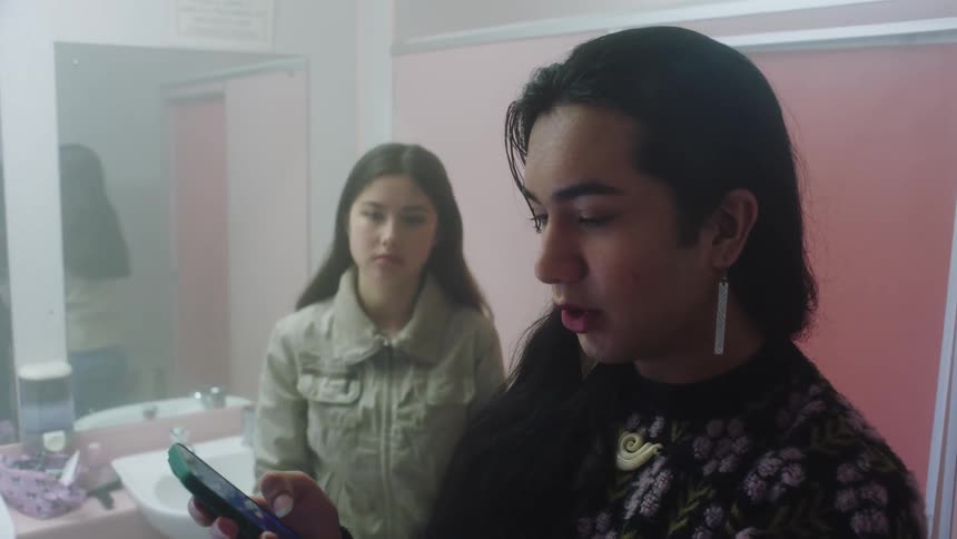 A woman talking on her cell phone while another makes up in the mirror and then goes to her cell phone too