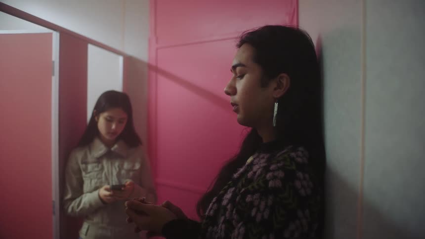 Two young ladies standing inside the restroom talking while leaning on the wall with one wearing a jacket and pressing her phone