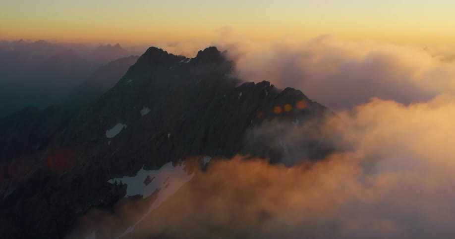 Drone view of dense fog rolling over high mountain peaks as the sun rises on the horizon
