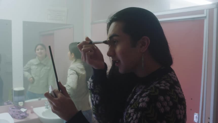 In the bathroom two women are present one woman is skillfully applying her makeup using a brush and holding a mirror in her hand while the other woman stands nearby attentively gazing at her reflection in the bathroom mirror