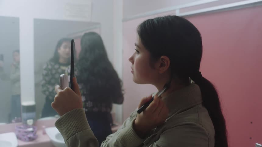 A woman running a makeup brush over her face while viewing herself in a hand mirror