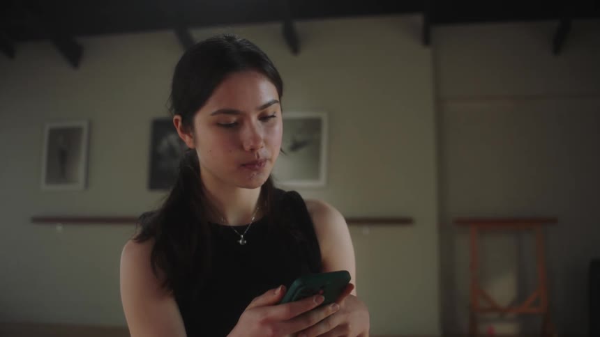 A woman seated in a room checking her phone and then picking up her water flask before standing up