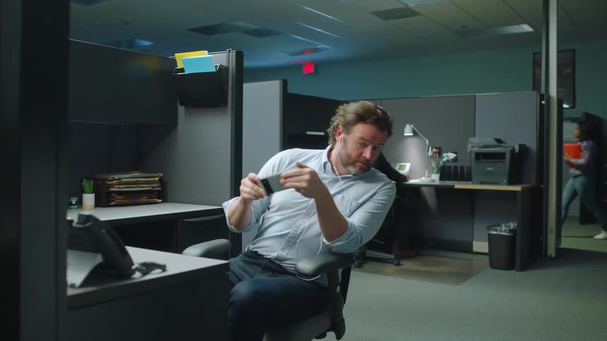 A man seated in the office holding his phone with both hands and then cautiously checks his surroundings by moving his chair to see if anyone is watching him