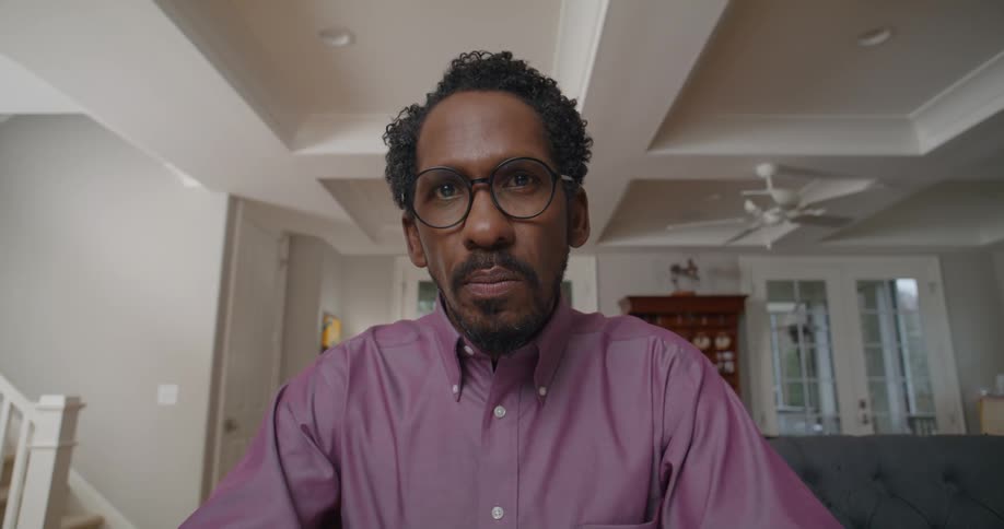A man in a long sleeve and glasses seated in a living room holding a pen engaging in a conversation