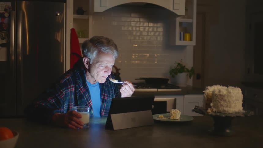 Man sitting in kitchen enjoys watching tablet while eating cake