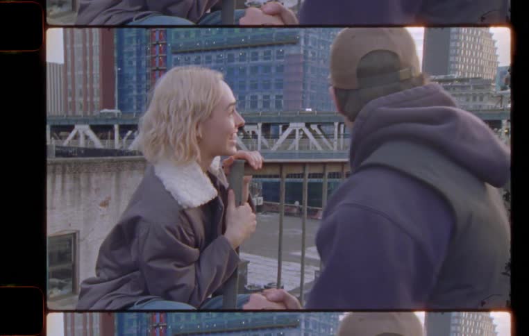 Man and woman climbs fire escape sits on building with bridge and skyline in the background at sunset