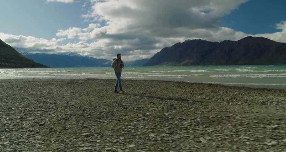 A man with a backpack is walking on a beach near the water with rocky cliffs in the background