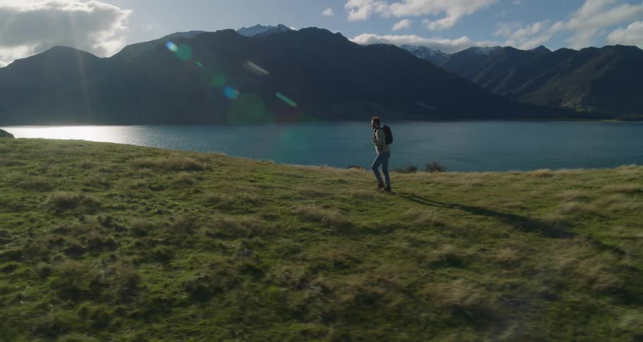 A man walking across a lawn in nature and in the background there is a huge lake and mountains