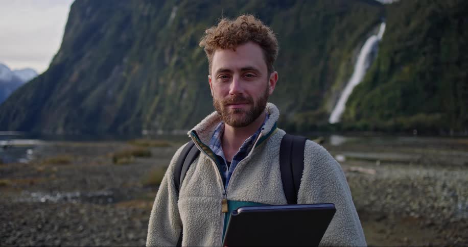Man with a happy facial expression holding a portable notebook and surrounded by mountains lake and waterfall