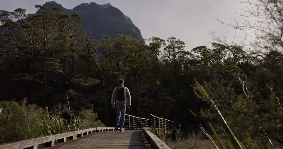 A man with a black backpack wearing a gray sweater and jeans walks across a bridge and into the forest with a high mountain in the background