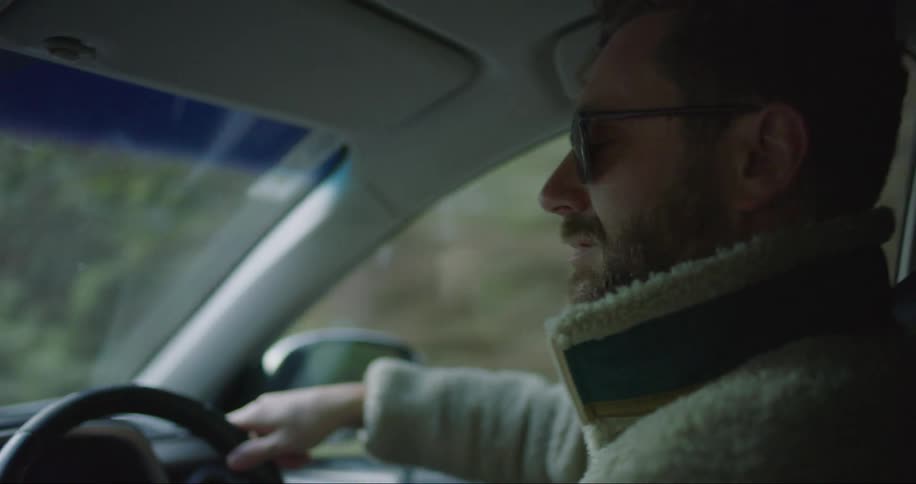 Close up of a man driving his car wearing sunglasses and a fuzzy sweater on a gloomy dau