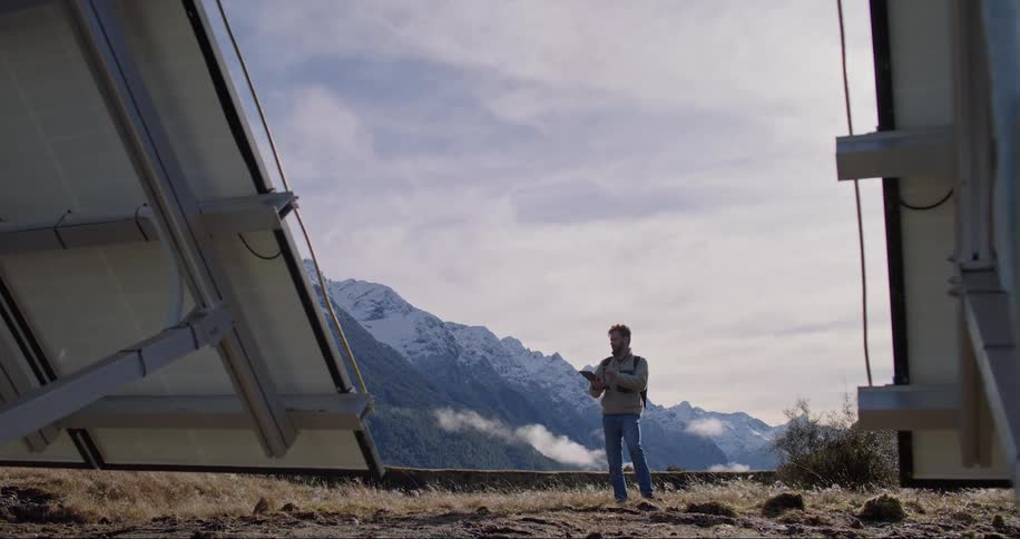 A man in a coat is fiddling with an electronic device while standing next to solar panels in the middle of nature