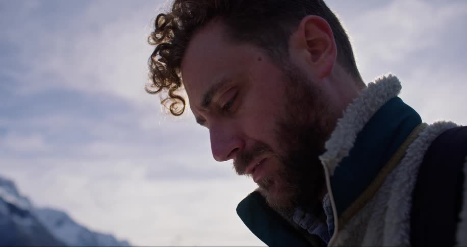A man standing outdoors near a mountain distressfully looks down and up and around under a blue clouded sky