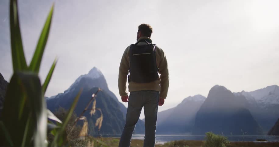 A man dressed in a gray sweater and jeans and carrying a backpack gazes at the high mountains in the distance