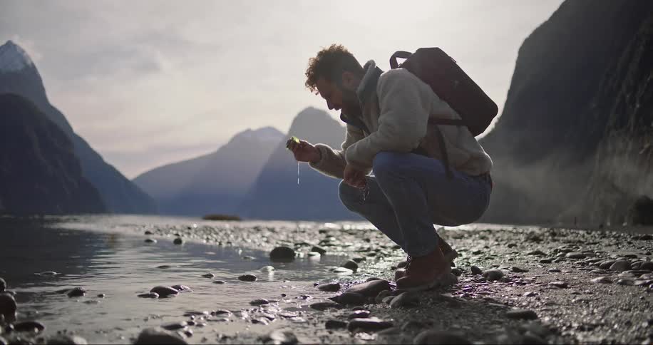 Hiker crouches near river and picks upitem to examine with mountains in the background