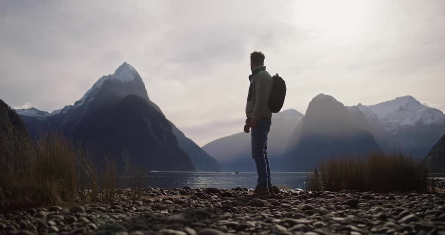 Man carrying a backpack stands on the shoreline of a lake in the mountains
