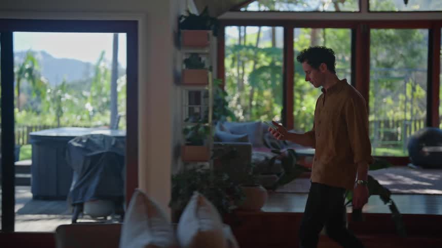 Man in brown shirt and black jeans walks across the living room in big house using smartphone