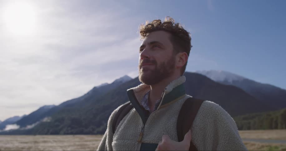 Young bearded man carrying a backpack stands on a valley near the mountains