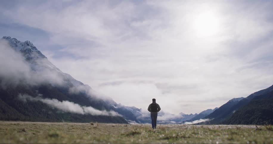 A man wearing a sweatshirt and backpack hikes through a grassy canyon between rocky cliffs