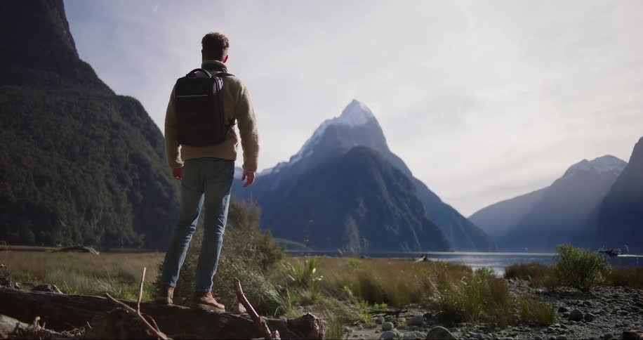 A man carrying a backpack walk and stand on a log of wood in the coastline covered in grass and small rocks with a boat see afar in the river surrounded by mountains