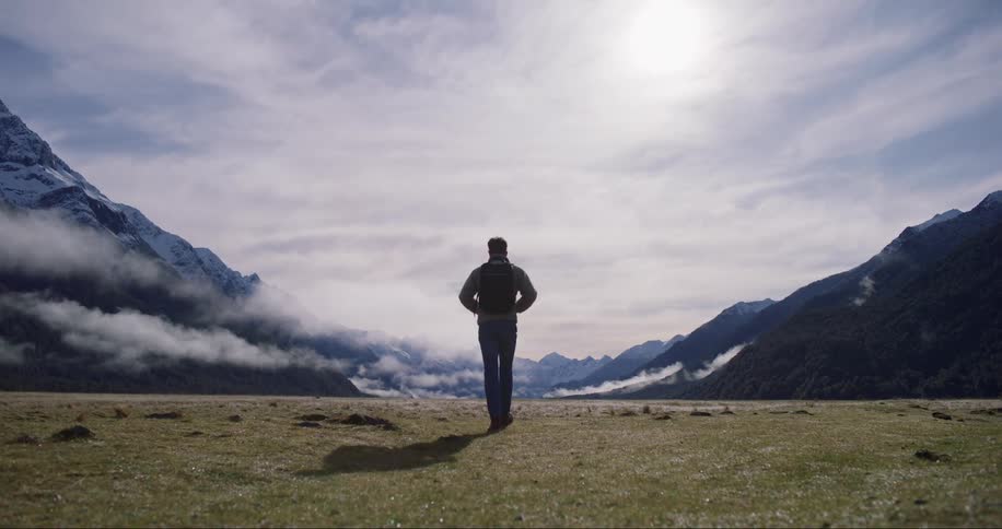 A man with a backpack walks toward a mountain range on a sunny day