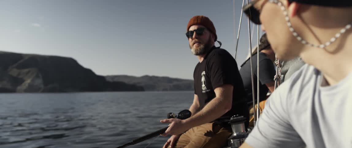 A man is fishing on a boat as he talks with a friend