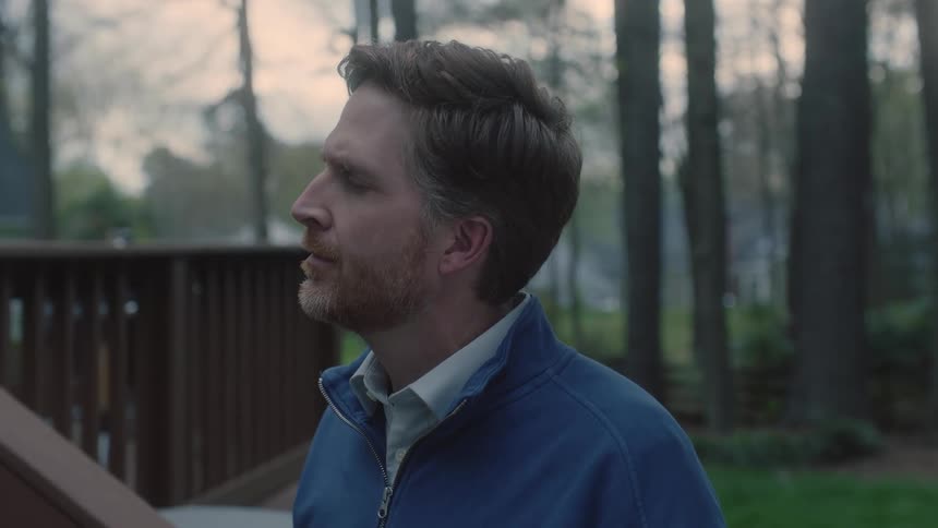 A beard man wearing a jacket standing on his backyard with lots of trees staring and contemplating