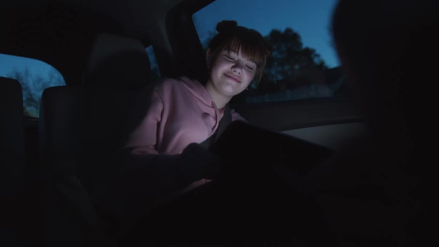 A young woman riding in a car looking at a tablet at night smiling and laughing