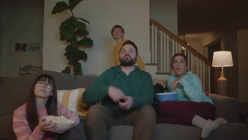 A man sitting on the sofa next to his wife and daughter eating popcorn with their teenage son passing by in the background
