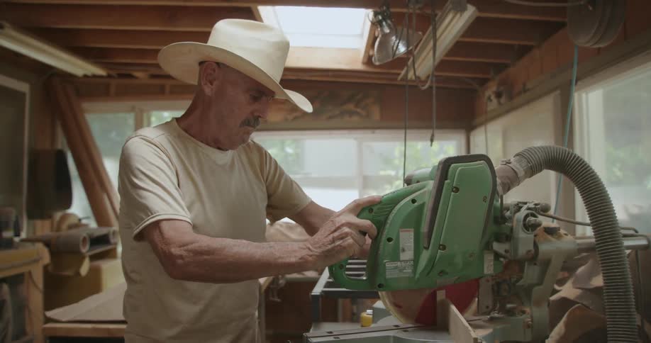 An older man wearing a white cowboy hat using a wood saw in his work shop during the day