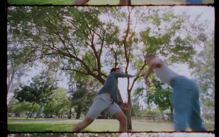 On a sunny day in the park under a tree two men are dancing ballet on the grass one is wearing jeans and a singlet while the other is wearing shorts and a shirt