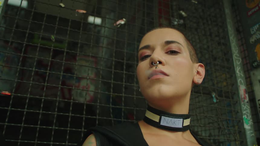 Close up of a woman wearing a nose ring and choker necklace while smoking a cigarette standing in front of a cage