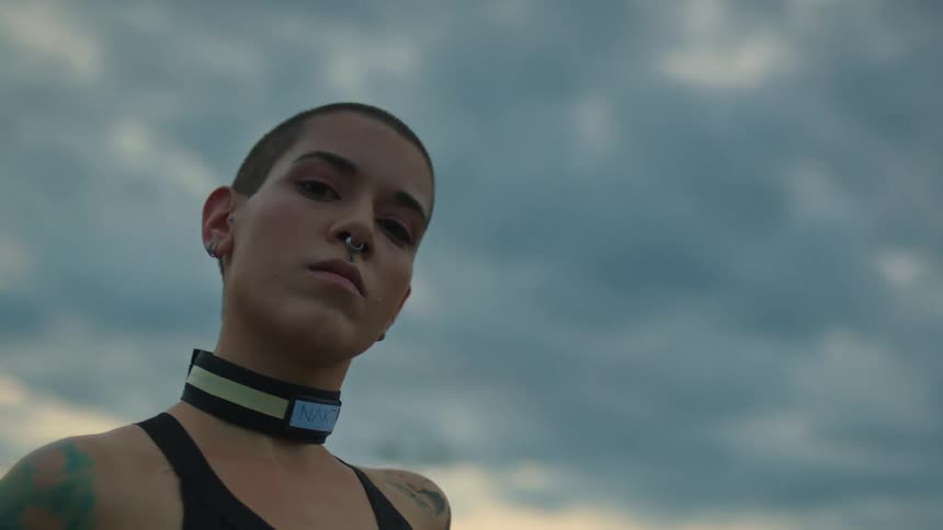 A woman standing outdoor while wearing a nose ring a belt on her neck with visible tattoo on her shoulder