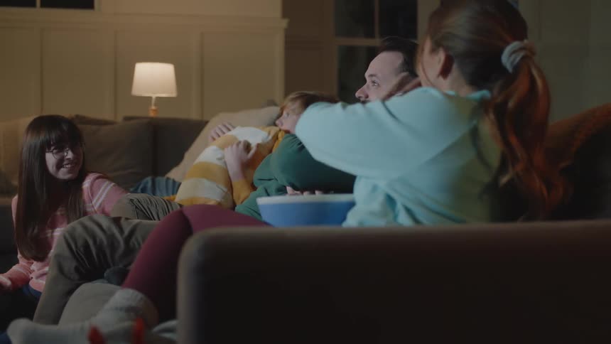A family sitting on a couch watching something on a bright screen and eating popcorn