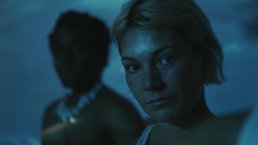 A woman staring in deep thought with someone behind in a dim lit room